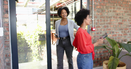 Entering two women talking while holding tablet and phone at office entry, glass door, red blouse. Duo, colleagues, collaboration, modern, entryway, sliding, brickの写真素材