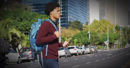 Walking man in maroon sweater, jeans with blue backpack, headset checking phone on road, copy space....