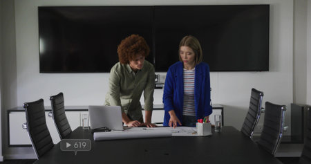 Checking two business architects examining blueprints on boardroom table silver laptop, markers. Collaboration, planning, architecture, design, schematics, teamwork, corporateの写真素材
