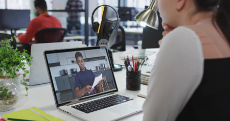 Watching remote presenter wearing dark top holding papers on laptop at office desk, lamp. Coworking, openplan, modern, daylight, monitors, collaboration, productivityの写真素材