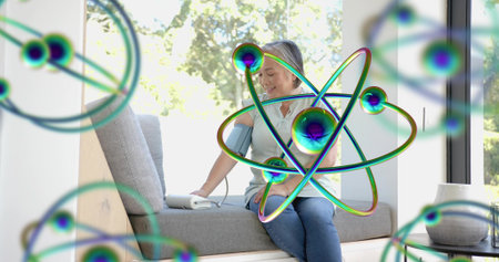 Sitting Asian woman checking BP cuff or monitor at window seat, light top, jeans. Bench, home,...