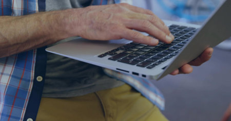 Typing mature man wearing plaid shirt and mustard pants, using slim silver laptop in lounge. Keyboard, notebook, casual, minimalist, interior, workspace, focusの写真素材
