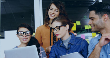 Collaborating team leaning over laptop and tablet at office in casual clothes, glass labeled CoLAB. Collaboration, brainstorming, ideation, coworkers, stickynotes, partition, modernの写真素材