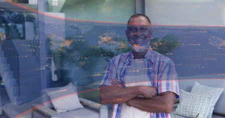 Standing mature man crossing arms facing camera in living room, in plaid shirt, glass reflections....