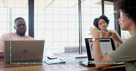 Gesturing woman in short-sleeve top leading meeting at wooden table glass office, laptops, tablet. Collaboration, boardroom, daylight, windows, discussion, charts, gridの写真素材