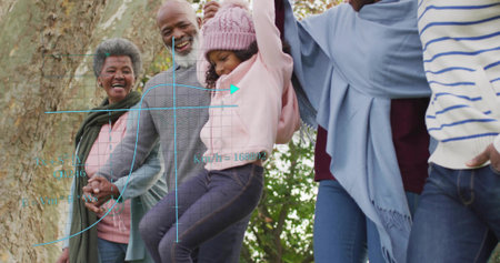 Lifting family holding hands, raising girl wearing knit hat at park near tree. Multigenerational, outdoors, trunk, foliage, autumn, warmth, togethernessの写真素材