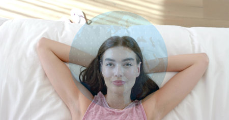 Reclining woman wearing pink sleeveless top on bed at home, with round pillow and globe overlay. Bedroom, relaxed, sunlit, sandals, cushion, orb, femaleの写真素材