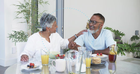 Holding hands couple wearing white blouse and blue T-shirt sharing breakfast at home, with pancakes. Partners, morning, minimalist, glass, table, plants, sunlitの写真素材