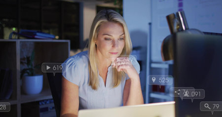 Working mature woman in light blue blouse using laptop at office, showing UI overlays. Desk, monitor, computer, lamp, plant, minimalist, analyticsの写真素材