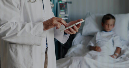 Standing Asian doc in lab coat tapping tablet, monitoring child in hospital bed, copy space. Doctor, medical, device, screen, room, clinical, careの写真素材