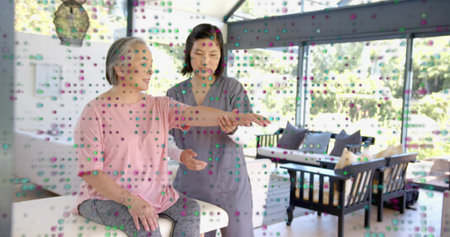 Caregiver wearing gray scrubs guiding senior in pink top and gray pants on ottoman, dotted glass. Therapy, eldercare, homecare, rehabilitation, mobility, assistance, wellnessの写真素材