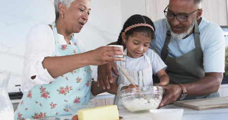 Stirring child wearing apron and pink headband, elders steadying glass bowl and pouring in kitchen. Family, multigenerational, baking, collaboration, home, bright, warmの写真素材