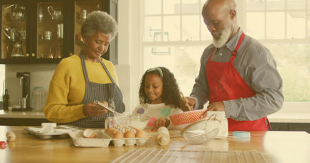 Stirring grandparents in aprons guiding granddaughter at kitchen island with mixing bowls and eggs....