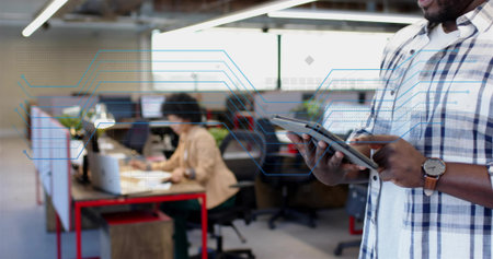 Standing adult man holding tablet and stylus in open-plan office, wearing plaid shirt, copy space. Coworking, collaboration, technology, monitors, productivity, modern, plantsの写真素材