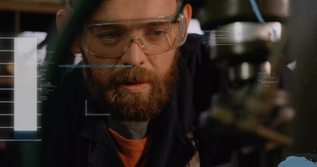 Bearded machinist inspecting spindle and chuck in workshop, wearing goggles, orange shirt with HUD....