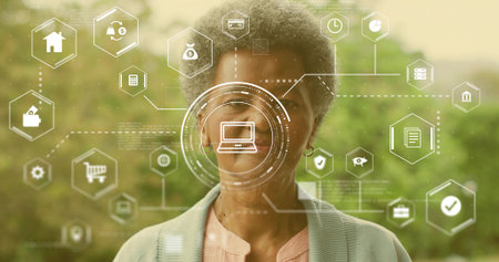 Standing senior woman wearing cardigan and pink blouse in garden, showing HUD laptop and hex icons....