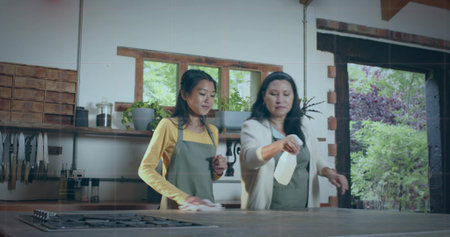 Cleaning two women wearing aprons wiping counter in rustic kitchen, with spray bottle and cloth....