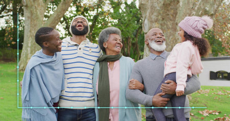 Smiling multigenerational family standing on grassy lawn under trees, wearing shawl and knit hat. Grandparents, child, park, autumn, outdoor, candid, cozyの写真素材
