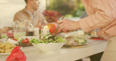 Tossing adult in pink cable-knit sweater using tongs, serving salad from white bowl at yard-table. Tableware, dishes, greens, alfresco, sunlit, warmth, candlelightの写真素材