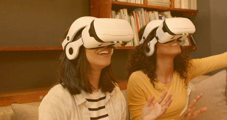 Reaching, smiling women wearing white VR headsets on sofa, striped and yellow tops near bookshelf....