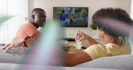 Sitting men in salmon-yellow tees on gray sofa at home watching mounted TV, holding clear bottle....