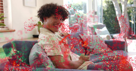 Smiling man wearing light shirt, shorts holding remote on blue sofa in living room, with confetti....