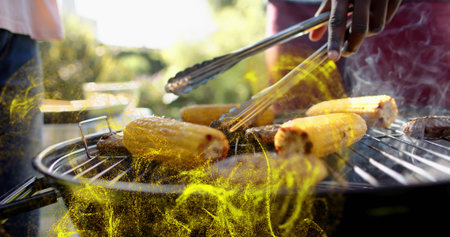 Turning metal tongs, men in shirts tending charcoal grill in backyard with corn and sausages, smoke. Barbecue, outdoor, cookout, food, summer, sunlit, casualの写真素材