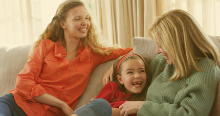 Laughing child red headband reclining between smiling women in orange shirt and green knit on sofa....