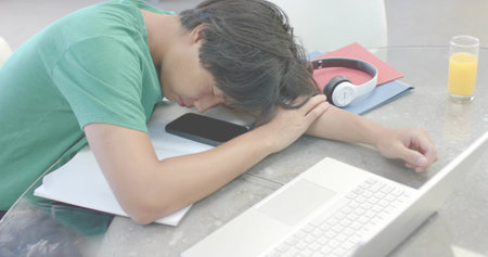 Napping Asian man in green tee leaning on glass table at home, with laptop, phone, headset. Sleepy, study, desk, headphones, papers, juice, brightの写真素材