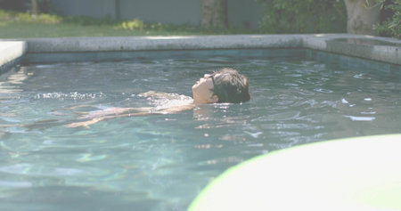 Floating adult Asian man reclining on back in residential pool, wearing sunglasses, near float....