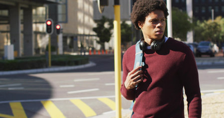 Walking man gripping light blue backpack strap at crosswalk, maroon sweater, headphones, copy space. Urban, daytime, sidewalk, commuter, streetstyle, sunlit, shadowsの写真素材