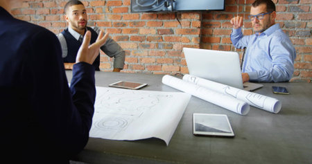 Reviewing lead wearing jacket gesturing to team in business around meeting table, plans and tablets. Architecture, design, collaboration, blueprints, industrial, brick, conferenceの写真素材