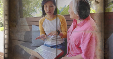 Showing raglan woman pointing as pink-shirt woman listening on porch bench with laptop, film-strip. Communication, intergenerational, mentorship, learning, collaboration, outdoor, greeneryの写真素材