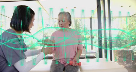 Therapist assisting woman in pink top, grey pants lifting pink dumbbells by glass walls, waveform. Caregiving, rehab, mobility, seated, exercise, senior, daylightの写真素材