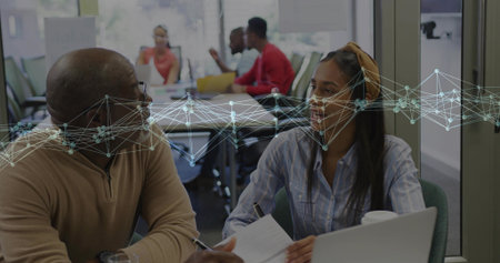 Discussing mentor in sweater and peer in blue shirt at meeting room with laptop papers overlay. Collaboration, teamwork, coworking, learning, daylight, windows, whiteboardの写真素材