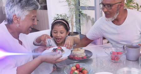 Smiling woman wearing white top offering plate with berries to child at home, pancakes on table....