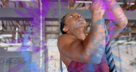 Climbing African American woman in red tank top gripping thick rope at gym, with paint-splash. Strength, fitness, focus, endurance, industrial, gritty, ascentの写真素材