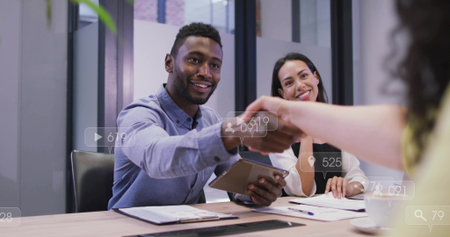 African American man in light-blue shirt holding tablet, shaking hands in conference room, UI icons. Boardroom, corporate, modern, collaboration, networking, professionalism, glassの写真素材