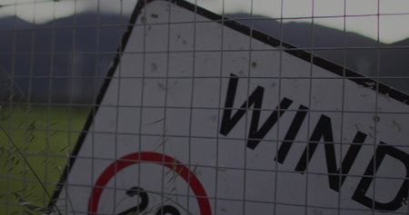 Showing tilted metal sign reading WIND behind wire mesh fence with tall grasses, red symbol. Weathered, warning, rustic, closeup, rural, overcast, fieldの写真素材