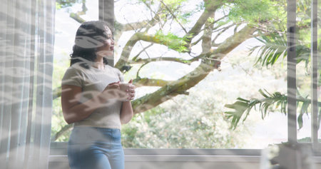 Holding white ceramic mug, woman wearing knit top and light blue jeans gazing out multi-pane window....