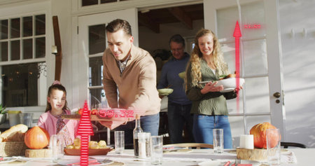 Passing family in sweaters and button-up placing platter at dining table in sunroom, with pumpkins....