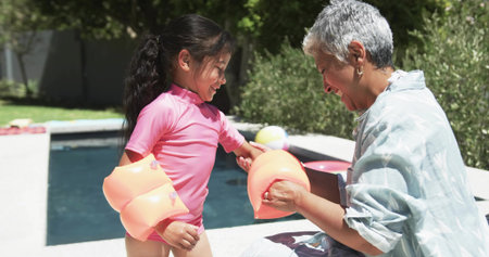 Adjusting senior woman fastening child's orange arm floatie at backyard pool edge, pink swim shirt. Grandparent, grandchild, outdoor, sunlit, waterplay, beachball, deckの写真素材