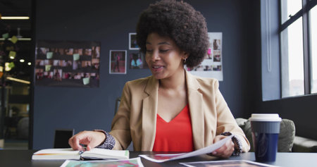 Reviewing printed pages, woman in tan blazer and red blouse sorting binder at meeting table. Office, studio, notebook, collage, window, daylight, coffeeの写真素材