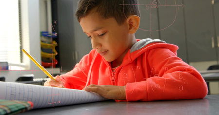 Writing child wearing red hoodie sitting at school desk, holding yellow pencil over spiral notebook. Classroom, student, study, learning, focus, concentration, binsの写真素材