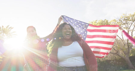 Laughing woman wearing white top and red cardigan standing in park, holding large American flag. Outdoors, backlit, lensflare, greenspace, trees, grass, happinessの写真素材
