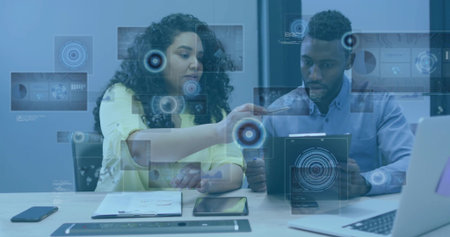 Pointing and explaining, pair in yellow blouse, light blue shirt holding tablet with AR graphics. Collaboration, teamwork, office, boardroom, table, touchscreen, dataの写真素材