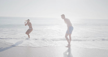 Jumping couple stepping back from incoming waves at shoreline, with bikini and blue swim trunks. Beach, ocean, backlit, highkey, windblown, reflection, coastlineの写真素材