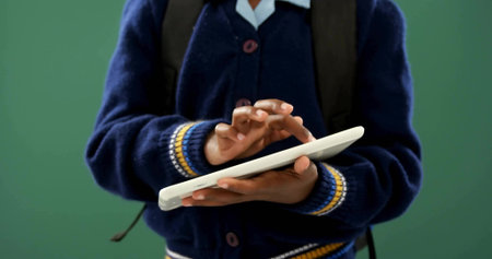 Tapping school student in navy cardigan and white collared shirt, holding white tablet and backpack. Education, girl, touchscreen, learning, edtech, classroom, greenの写真素材