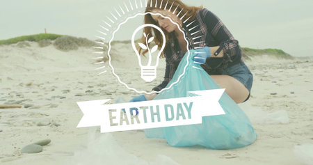 Kneeling woman collecting trash on beach, blue-gloved, plaid, holding blue bag, Earth Day overlay. Shore, litter, cleanup, outdoors, dunes, overcast, volunteerの写真素材