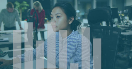 Working Asian woman wearing striped top, focusing on monitor in night office with bar-chart overlay....